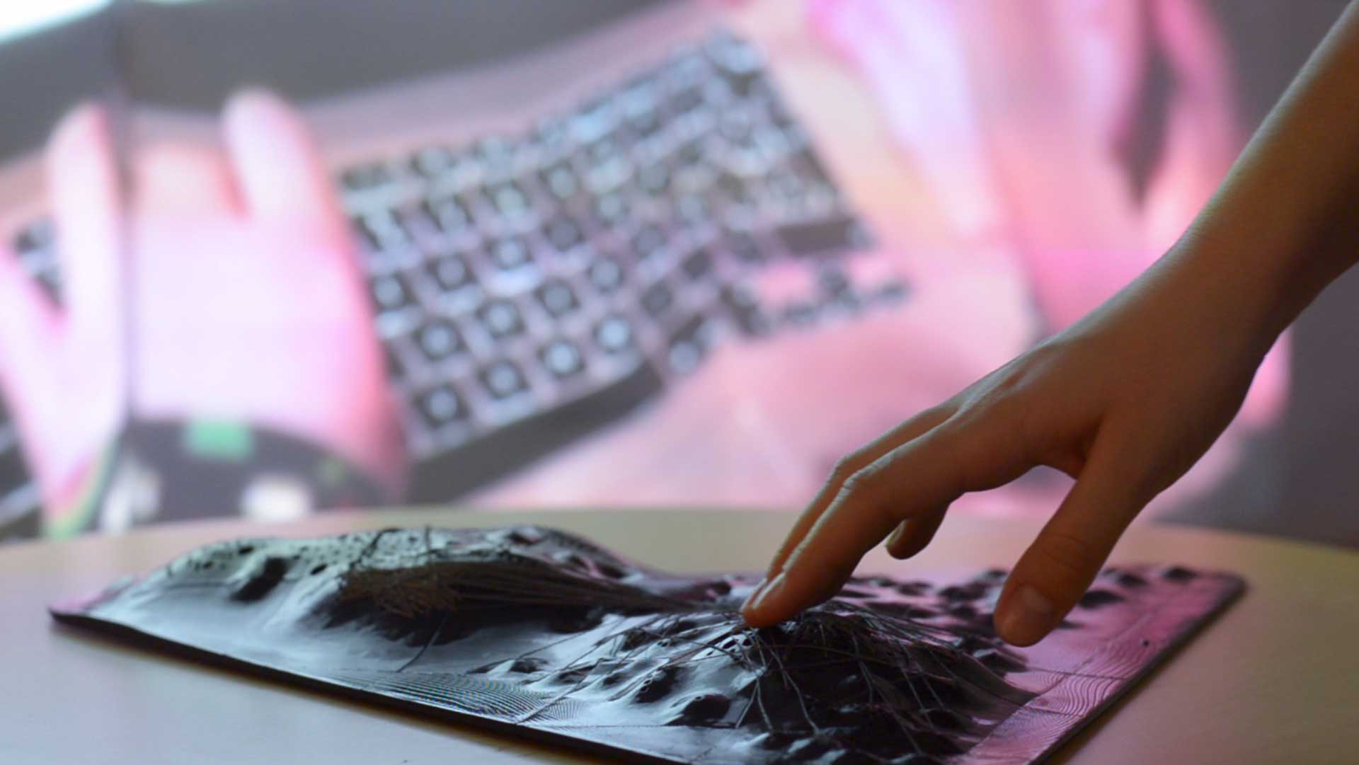 In the foreground is a hand touching the artwork, a topographic object with conductive threads, in the background is a projection on a wall of a hand touching a laptop keyboard.