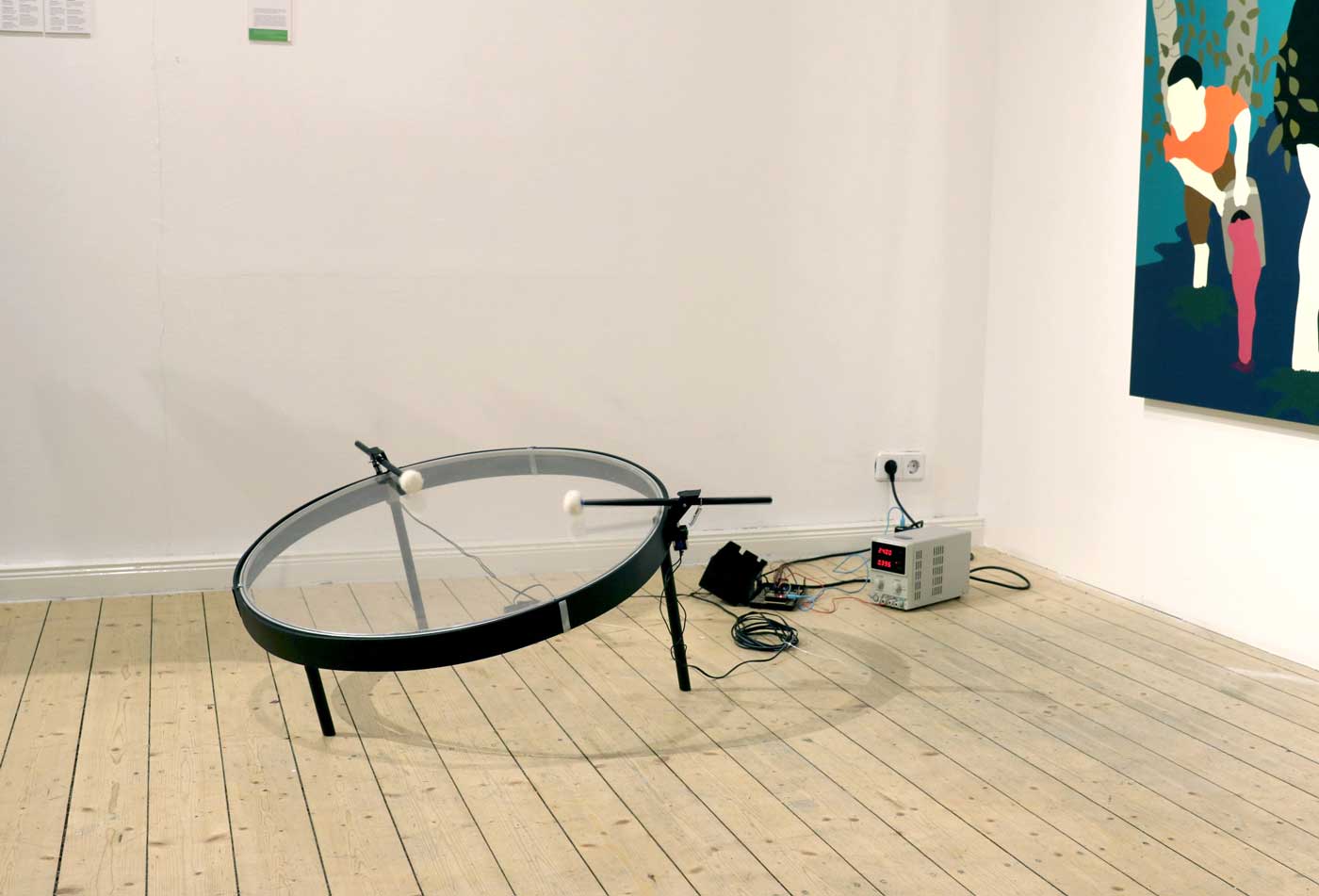An exhibition photo, an electrical power supply is connect to a power socket, indistinguishable electronics, and a large transparent drum frame being struck by mallets.