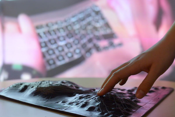 In the foreground is a hand touching the artwork, a topographic object with conductive threads, in the background is a projection on a wall of a hand touching a laptop keyboard.